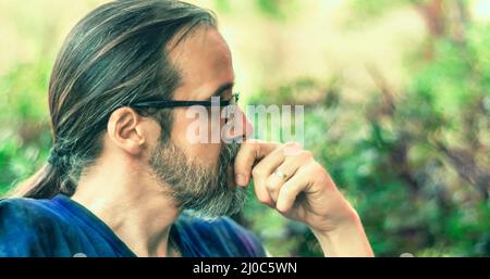 Portrait of one mid adult man with beard, glasses and long hair in a ponytail, looking thoughtful. Side view. Selective focus Stock Photo