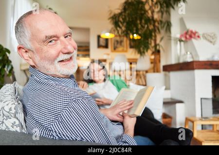 Happy retired senior man reading book in living room with wife on background Stock Photo