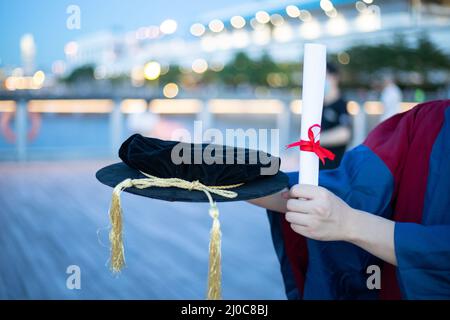 happy proud PhD graduated male student in Academic dress gown holds Graduation cap or hat and diploma or certificate. Blurred night colorful city ligh Stock Photo