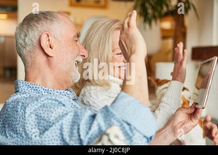 Senior couple waving happily while video chatting with family on tablet computer at home Stock Photo