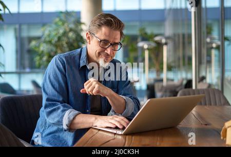 Happy middle age businessman using laptop computer in office. Older casual man working online on terrace of cafe, smiling. Stock Photo