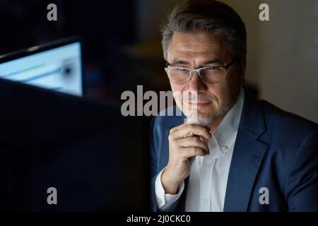 Businessman working with computer at office desk, thinking, smiling. Portrait of happy mid adult man in shirt and jacket, smiling. Bearded, glasses. Stock Photo