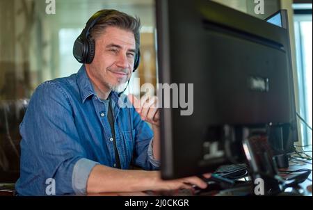 Businessman working with computer in office talking on headset. Man sitting at desk in front of screen, smiling, happy. Customer service support operator Stock Photo
