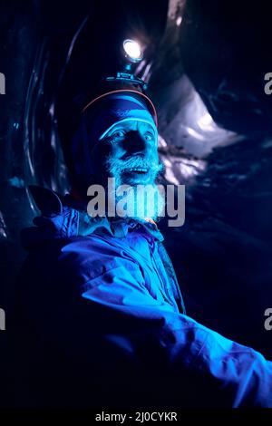 Portrait of Old Alpinist with grey beard in helmet with glow headlamp smiling in deep dark glacial ice cave with blue lights explore winter mountain g Stock Photo