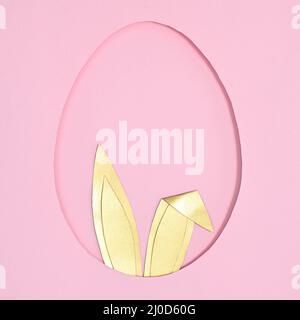 Egg shape frame with golden rabbit ears peek from hole on pastel pink background. Flat lay creative Easter concept Stock Photo