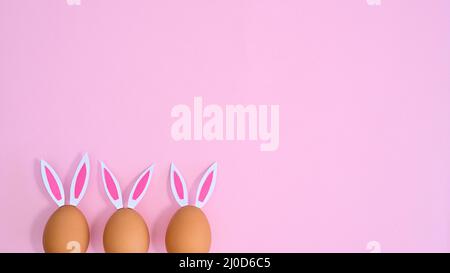 Three natural eggs with rabbit ears peek on pastel pink background with copy space. Easter flat lay minimal Stock Photo