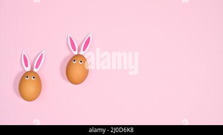 Two natural eggs with bunny ears and eyes on pastel pink background. Flat lay copy space Easter concept Stock Photo
