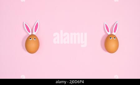 Two natural eggs with bunny ears and eyes on pastel pink background. Flat lay copy space Easter concept Stock Photo