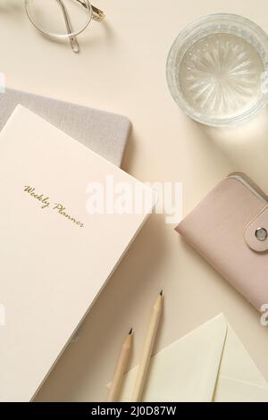 Feminine home office desk with weekly planner, glass of water, eyeglasses, pencils on beige table. Stock Photo