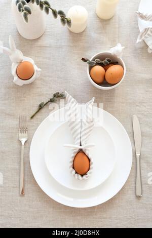 Easter table setting with egg in Easter bunny napkin on plate, fork and knife, willow on linen tablecloth. Stock Photo