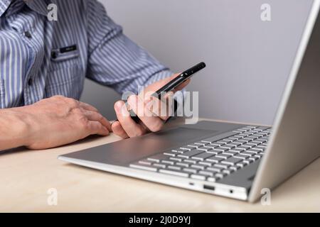 Man sitting at table with laptop and holding phone. Checking information, reading or typing message while working on computer. Man hands closeup. High quality photo Stock Photo