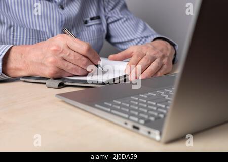 Man taking notes in planner and using laptop for online education, distant work. Hands closeup. Man writing schedule, thoughts, important information. High quality photo Stock Photo
