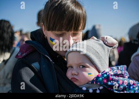 Warsaw, Poland. 18th Mar, 2022. A Ukrainian mother holds her baby with Ukrainian flag painted on her cheek during the protest. The 'March of Ukrainian Mothers' was held in Warsaw, which was also a protest against the war and killing of Ukrainian children. The slogans of the march were 'World, help our children', 'Stop the war', 'Save kids of Ukraine' and 'Close the sky'. Mainly Ukrainian mothers and their children, who found refuge in Poland from the war, took part in the protest. Credit: SOPA Images Limited/Alamy Live News Stock Photo