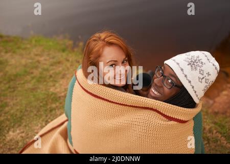 As close as two best friends can get. A high angle portrait of two happy young women huddled under a blanket next to a lake in winter. Stock Photo