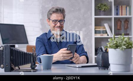 Bearded man working online with laptop computer and smart phone at home sitting at desk. Businessman in home office. Portrait of mature age, middle age, mid adult man in 50s. Stock Photo