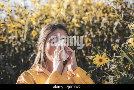 Cold flu season, runny nose. Flowering trees in background. Young girl sneezing and holding paper tissue in one hand and flower bouquet in other. Flu Stock Photo
