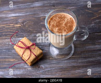 Spicy warming tea with milk in a glass cup with cinnamon, gingerbread cookie tied with ribbon and Christmas tree branches on a wooden table, rustic st Stock Photo