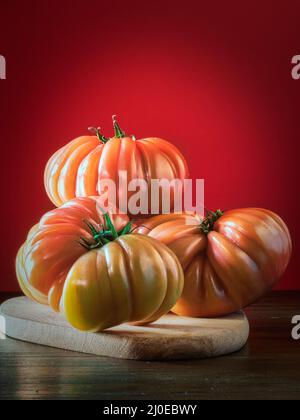 Close-up of three ripe tomatoes placed on a wooden board. Red background. Beefsteak tomatoes. Stock Photo