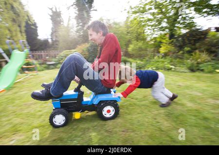 Son pushing his father on a toy tractor in their back garden themes of exhilaration family bonding Stock Photo