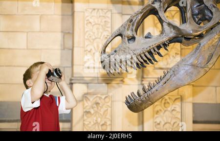 A caucasian young boy photographing a Tyrannosaurus rex dinosaur with an old camera in a museum Stock Photo