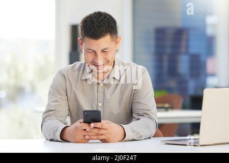 I need to check-in with a client. Shot of a handsome young businessman sitting alone in his office and using his cellphone during the day. Stock Photo
