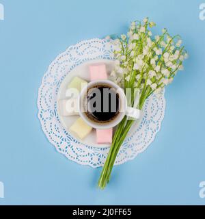 White cup of coffee latte on a saucer, marshmallow, a bouquet of lily of the valley flowers on blue Stock Photo