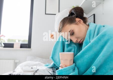 A young teenager drinks hot tea with herbs while lying on her bed under the covers. Stock Photo