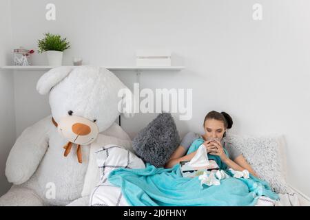 A young teenager drinks hot tea with herbs while lying on her bed under the covers. Stock Photo