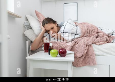 On the bed lies a teenager and in front of her stands a table with apples and medicines that must be swallowed. Stock Photo