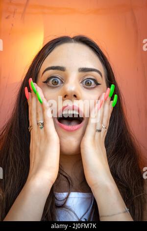 Shocked or surprised beautiful young woman with colored nails holding her cheeks  and shouting Stock Photo