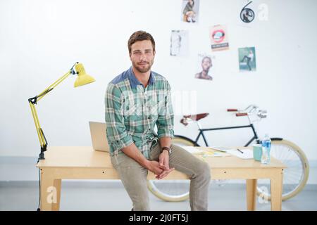Portrait of a casually-dressed young man sitting on his desk in an office. The commercial designs displayed in this image represent a simulation of a Stock Photo