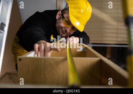 A construction worker checks a pull-out cupboard at the top with a tape measure. Wearing personal protective equipment. Experien Stock Photo