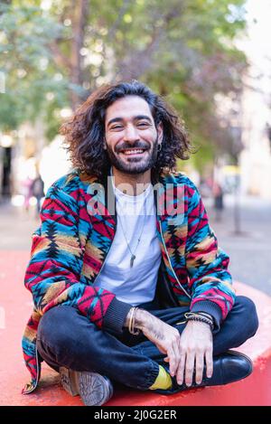 Optimistic Hispanic male in stylish clothes with beard and dark hair smiling. He is looking at camera while sitting cross legged on bench on sunny day in park Stock Photo