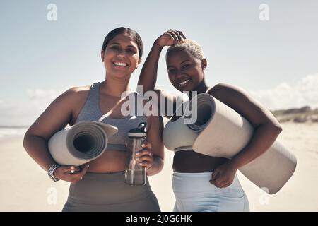 Friends who sweat together, stick together. Shot of two young women holding their yoga mats while on the beach. Stock Photo