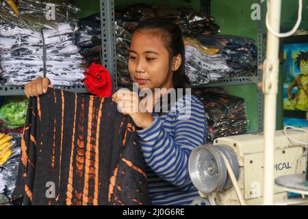 Young female dressmaker or fashion designer sitting near her sewing machine with new beautiful batik dress in hands. Focused woman owner of local Stock Photo