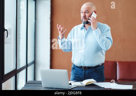 Serious mature businessman talking by phone staying by table in office Stock Photo