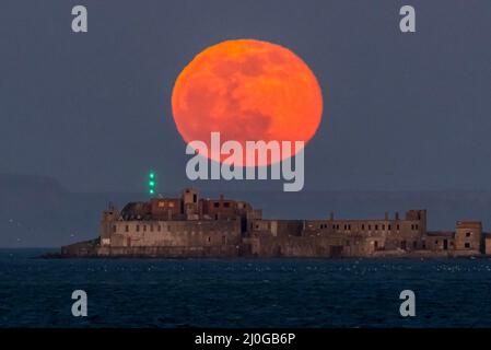 Weymouth, Dorset, UK.  18th March 2022.  UK Weather.  The full Worm Moon glows red as it rises up from behind the historic 19th century Portland Breakwater Fort on the outer breakwater of Portland Harbour near Weymouth in Dorset.  The now derelict fort also known as Chequered Fort was constructed between 1868 and 1878 and is Grade II listed.  Picture Credit: Graham Hunt/Alamy Live News