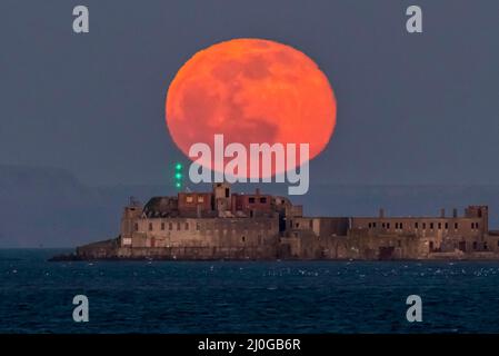 Weymouth, Dorset, UK.  18th March 2022.  UK Weather.  The full Worm Moon glows red as it rises up from behind the historic 19th century Portland Breakwater Fort on the outer breakwater of Portland Harbour near Weymouth in Dorset.  The now derelict fort also known as Chequered Fort was constructed between 1868 and 1878 and is Grade II listed.  Picture Credit: Graham Hunt/Alamy Live News