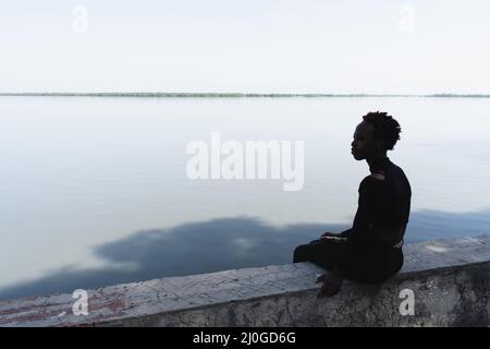 Silhouette of a lonely young African teenage girl sitting on a wall by the river and looking at the other bank; symbols of solitude, longing, nostalgi Stock Photo