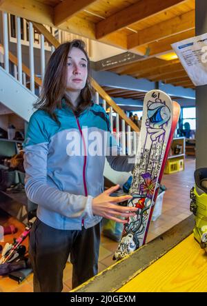 Woman with brown hair looking, renting ski equipment, holding her ski while waiting for her helmet and shoes, standing in side a building, staircase b Stock Photo
