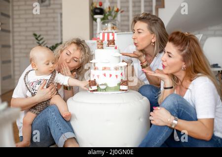 Three attractive middle-aged and young women blowing kisses to little one-year-old boy touching bear on birthday cake. Stock Photo