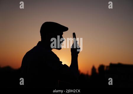 Silhouette portrait of man investigator in peaking cap looking with magnifying at side. Man searching sales or discounts at sunset using loupe with sun on background. High quality image Stock Photo