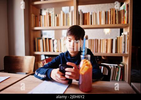 Boy rests after school, teenager sit in cafe and drink juice coctail, watching on mobile phone. Stock Photo