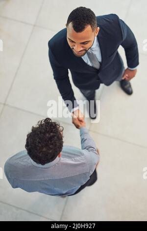 Thanks for stopping by. High angle shot of two confident young businessmen shaking hands in agreement while standing in the office during the day. Stock Photo
