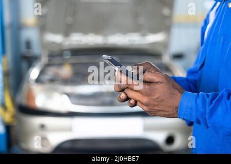 Close up of mechanic hands busy using mobile phone in front of broken car at garage or workshop - concept of communication, internet and technology Stock Photo