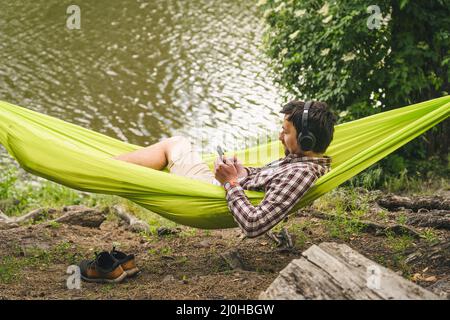 Outdoors and adventure concept. Bike trip to forest. Cyclist is resting in green hammock between trees in nature by lake while l Stock Photo