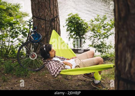 Outdoors and adventure concept. Bike trip to forest. Cyclist is resting in green hammock between trees in nature by lake while l Stock Photo