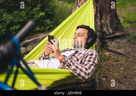 Outdoors and adventure concept. Bike trip to forest. Cyclist is resting in green hammock between trees in nature by lake while l Stock Photo
