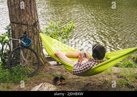 Outdoors and adventure concept. Bike trip to forest. Cyclist is resting in green hammock between trees in nature by lake while l Stock Photo