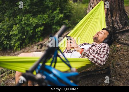 Young caucasian man resting in hammock, listening to music on headphones and using smartphone afterwards on bicycle in forest ne Stock Photo
